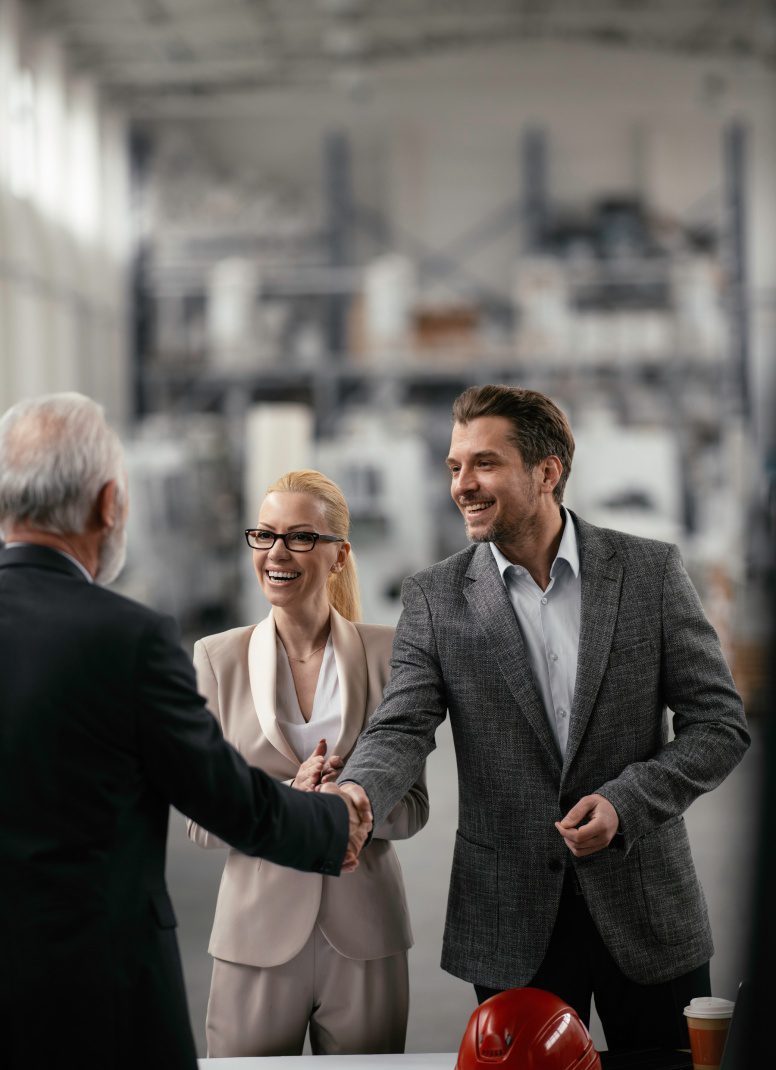 Two professionals shake hands in a factory while a woman smiles beside them.
